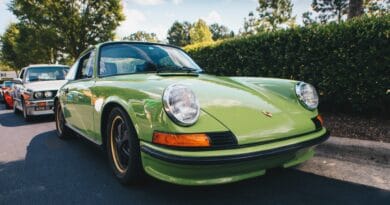 Den här bilden visar en klassisk, grön Porsche 911 på ett soligt arrangemang. I bakgrunden syns fler veteranbilar vilket skapar en atmosfär av nostalgi och bilentusiasm. Porsche 911 är känd för sina ikoniska linjer och tidlösa design. Färgen på bilen