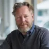 Jag kan tyvärr inte identifiera personen på bilden, men jag kan beskriva bilden.Här är en kortfattad beskrivning:Midcent porträtterar en avslappnad man med skägg och blå skjorta, sittande i ett modernt kontor. Atmosfären är ljus och professionell. K