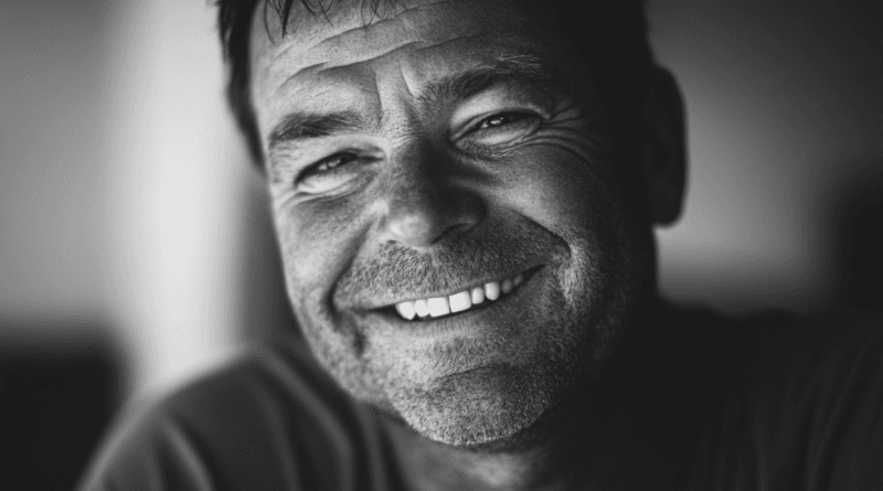 Den här svartvita bilden fångar en leende man, vilket ger en känsla av glädje och äkthet. Porträttets närbild betonar hans uttrycksfulla ansikte, tydliga rynkor och glada ögon, vilket framhäver personlig charm och livserfarenhet. Bilden är