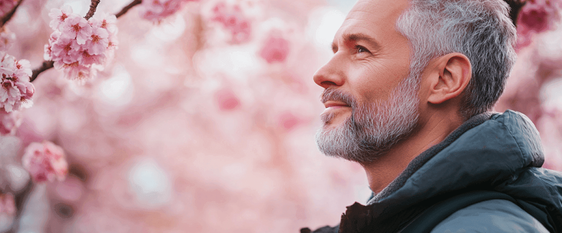 Midcent: En man med grått hår och skägg njuter av våren bland blommande körsbärsträd, med kamera i handen. Harmonisk naturupplevelse i fokus. Resa.