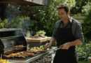En man i grå skjorta och förkläde står vid en grill utomhus, omgiven av grönska. Han hanterar grillverktyg och olika livsmedel tillagas. Midcent Mat.