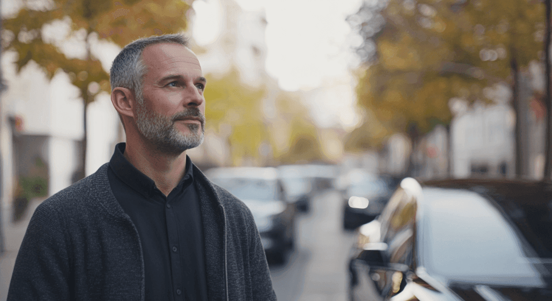 En man står avslappnad på en gata kantad av träd i höstens färger. Han ser nöjd och eftertänksam ut, vilket skapar en reflekterande atmosfär. Midcent - Samhälle.