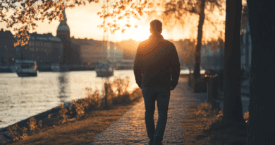 Midcent: En man promenerar längs en strandpromenad vid solnedgången, omgiven av vatten och båtar. Solens gyllene strålar skapar en lugn och reflekterande atmosfär, perfekt för en rofylld stund. Kategori: Resa.