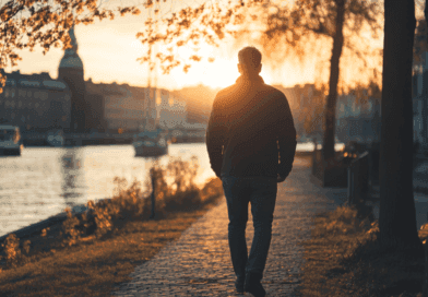 Midcent: En man promenerar längs en strandpromenad vid solnedgången, omgiven av vatten och båtar. Solens gyllene strålar skapar en lugn och reflekterande atmosfär, perfekt för en rofylld stund. Kategori: Resa.