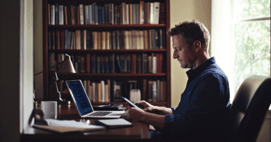 Midcent: En man fokuserar på sin mobiltelefon och laptop i ett hemtrevligt kontor omgivet av böcker. Bilden förmedlar lugn och produktivitet. Perfekt för arbete eller studier. Kategori: Teknik.