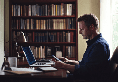 Midcent: En man fokuserar på sin mobiltelefon och laptop i ett hemtrevligt kontor omgivet av böcker. Bilden förmedlar lugn och produktivitet. Perfekt för arbete eller studier. Kategori: Teknik.