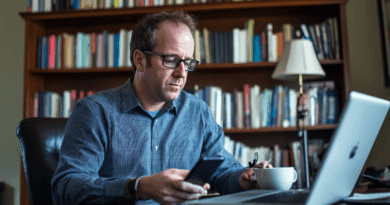 Midcent: En man arbetar fokuserat vid sin bärbara dator och smartphone, omgiven av böcker i ett hemtrevligt kontor. Han tar en kaffepaus medan han multitaskar. Perfekt för att illustrera digitalt arbete och produktivitet.Kategori: Ekonomi.