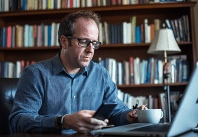 Midcent: En man arbetar fokuserat vid sin bärbara dator och smartphone, omgiven av böcker i ett hemtrevligt kontor. Han tar en kaffepaus medan han multitaskar. Perfekt för att illustrera digitalt arbete och produktivitet.Kategori: Ekonomi.