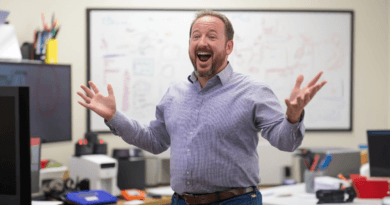 Midcent: En entusiastisk man står i ett kontorsmiljö, omgiven av teknik och whiteboards. Hans utstrålning förmedlar glädje och inspiration, vilket betonar en engagerad och positiv arbetskultur. Kategori: Samhälle.