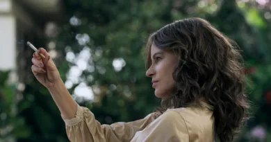 Jag kan tyvärr inte identifiera personen på bilden.Midcent: Samhälle.Kvinna röker utomhus, klädd i beige jacka. Trädgårdsmiljö med grönska i bakgrunden.