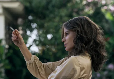 Jag kan tyvärr inte identifiera personen på bilden.Midcent: Samhälle.Kvinna röker utomhus, klädd i beige jacka. Trädgårdsmiljö med grönska i bakgrunden.