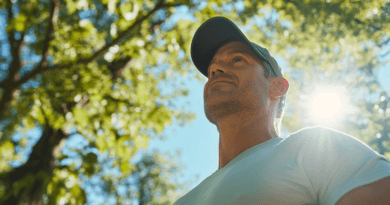 En medelålders man under en solig dag i naturen, där grönska och solljus skapar en harmonisk atmosfär. Bilden fångar känslan av att vara ute i det fria. Midcent – Hälsa.