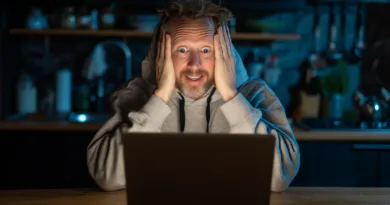 Midcent – En medelålders man sitter fascinerad framför en laptop i ett mörkt kök med ett överraskat uttryck. Perfekt för att illustrera fascination och teknikens förmåga att fängsla. Kategori: Underhållning.