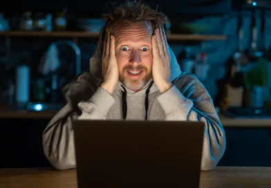 Midcent – En medelålders man sitter fascinerad framför en laptop i ett mörkt kök med ett överraskat uttryck. Perfekt för att illustrera fascination och teknikens förmåga att fängsla. Kategori: Underhållning.