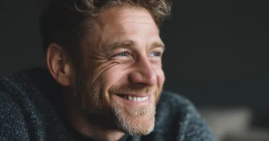 Midcent presenterar en medelålders man med ett varmt leende, vilket speglar glädje och optimism. Bilden fångar en avslappnad och naturlig atmosfär. Han bär en bekväm tröja och har kort, lockigt hår och skägg. Kategori: Samh