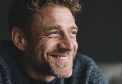 Midcent presenterar en medelålders man med ett varmt leende, vilket speglar glädje och optimism. Bilden fångar en avslappnad och naturlig atmosfär. Han bär en bekväm tröja och har kort, lockigt hår och skägg. Kategori: Samh