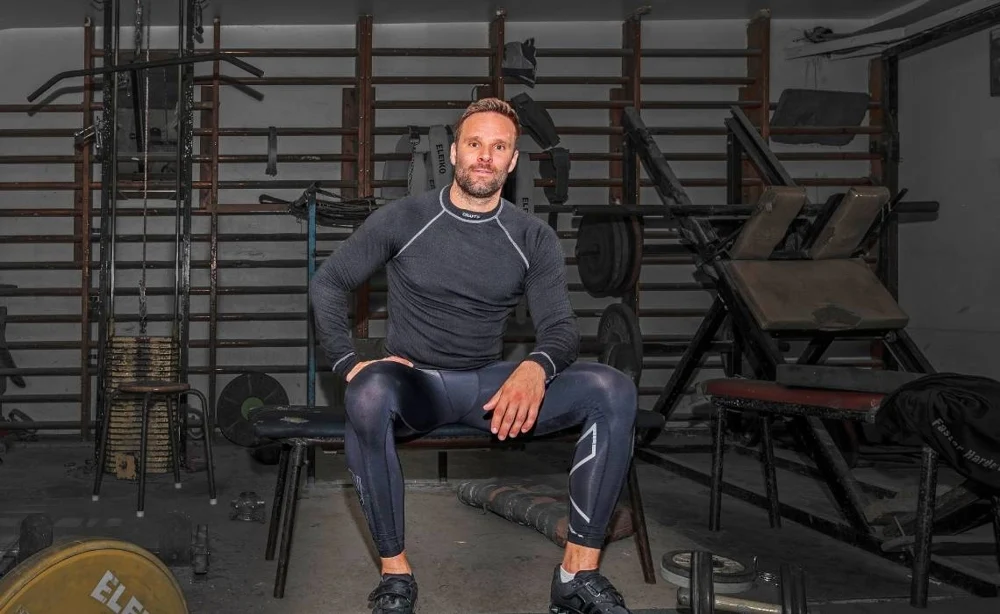 Midcent: En medelålders man i träningskläder sitter på en bänk i ett gym, omgiven av träningsutrustning och vikter. Bilden fångar en hälsosam och aktiv livsstil. Kategori: Hälsa.