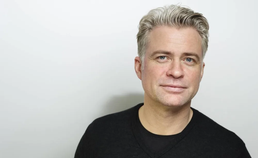 Jag kan tyvärr inte identifiera personen på bilden. Här är en beskrivning:En medelålders man med grått hår och svart tröja mot vit bakgrund, leende. Midcent. Kategori: Samhälle.