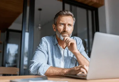 Midcent: En medelålders man sitter vid en dator, djupt försjunken i tankar. Han bär en randig skjorta och betraktar skärmen i ett modernt kontor eller hem. Perfekt för teman inom arbete och fokus. Kategori: Ekonomi.