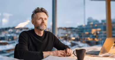 AI-hysterin: Varför börjar både svenskar och företag få kalla fötter? Midcent: En medelålders man reflekterar vid ett kontorsbord med laptop och kaffekopp, omgiven av en stadsutsikt. Arbetsmiljö och kreativitet står i fokus. Kategori: Ekonomi.