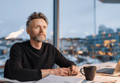 Midcent: En medelålders man reflekterar vid ett kontorsbord med laptop och kaffekopp, omgiven av en stadsutsikt. Arbetsmiljö och kreativitet står i fokus. Kategori: Ekonomi.