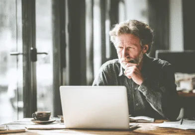 En medelålders man arbetar fokuserat vid en dator i ett stilfullt kontor. Han dricker kaffe medan han går igenom viktiga dokument. Bilden förmedlar en atmosfär av professionalism och koncentration.Midcent: Ekonomi.