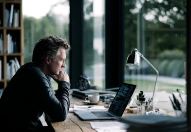 Midcent: En medelålders man arbetar fokuserat vid en bärbar dator i ett modernt hemmakontor. Fönstren släpper in naturligt ljus med utsikt över natur. Träbord med diverse kontorsmateriel. Teknik.