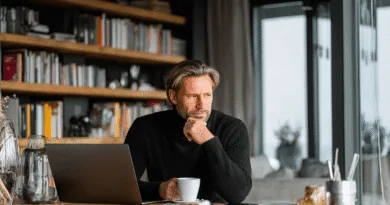 Midcent: En medelålders man sitter vid ett skrivbord med en laptop, omgiven av böcker och håller en kaffekopp, försjunken i tankar. Den hemtrevliga miljön och koncentrationen antyder arbetsro. Kategori: Samhälle.