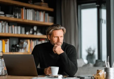 Midcent: En medelålders man sitter vid ett skrivbord med en laptop, omgiven av böcker och håller en kaffekopp, försjunken i tankar. Den hemtrevliga miljön och koncentrationen antyder arbetsro. Kategori: Samhälle.