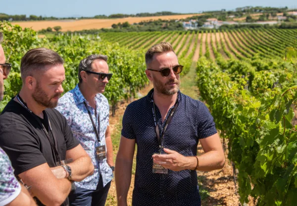 En medelålders man guidar en grupp genom en grönskande vingård under en solig dag. Omgivna av frodiga vinrankor, diskuteras troligen vinodling och produktionstekniker. Midcent. Kategori: Mat.