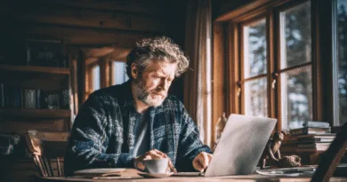 Midcent: En medelålders man arbetar fokuserat vid en bärbar dator i ett mysigt träinteriör, omgiven av böcker. Fönstren släpper in naturligt ljus och en kaffekopp står bredvid. Kategori: Samhälle.