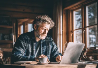 Midcent: En medelålders man arbetar fokuserat vid en bärbar dator i ett mysigt träinteriör, omgiven av böcker. Fönstren släpper in naturligt ljus och en kaffekopp står bredvid. Kategori: Samhälle.