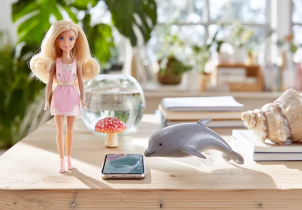 Docka i rosa klänning på bord med smartphone, fiskboll, och delfinfigur i soligt rum. Passar barnleksaker och inredningsdetaljer. Midcent. Underhållning.