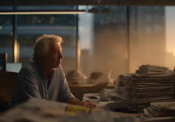 Midcent: En medelålders man arbetar sent på sitt kontor, omgiven av tidningshögar och datorer i kvällsljus. Scenen förmedlar