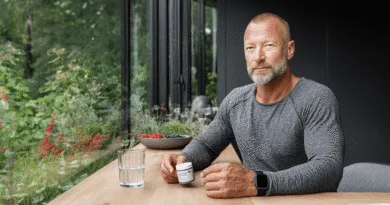 En medelålders man sitter vid ett bord bredvid ett fönster. Han håller en burk med kosttillskott. I bakgrunden syns grönska och en skål med bär. Han bär en grå tröja och en smartklocka. Midcent. Hälsa.