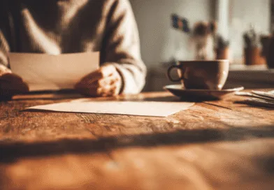 Midcent: En medelålders man sitter vid ett rustikt träbord i varmt ljus och läser en brev eller dokument. I förgrunden ligger papper, bredvid en kaffekopp. Avslappnad atmosfär dominerar bilden. Kategori: Samhälle.