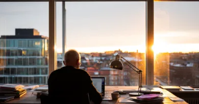 Midcent: En medelålders man arbetar koncentrerat vid sitt skrivbord med utsikt över en stad i solnedgång. Många papper ligger runtomkring, vilket ger en känsla av kreativitet och fokus. Kategori: Samhälle.