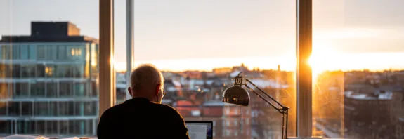 Midcent: En medelålders man arbetar koncentrerat vid sitt skrivbord med utsikt över en stad i solnedgång. Många papper ligger runtomkring, vilket ger en känsla av kreativitet och fokus. Kategori: Samhälle.
