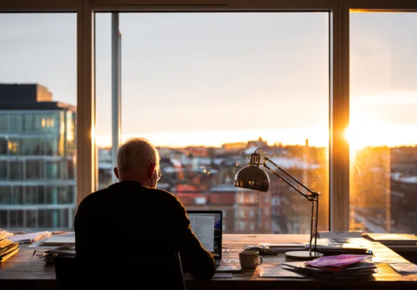 Midcent: En medelålders man arbetar koncentrerat vid sitt skrivbord med utsikt över en stad i solnedgång. Många papper ligger runtomkring, vilket ger en känsla av kreativitet och fokus. Kategori: Samhälle.