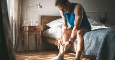 En medelålders man sitter på sängkanten och gnuggar sitt ben i morgonsolens sken, vilket indikerar smärta eller stelhet. Midcent. Hälsa.