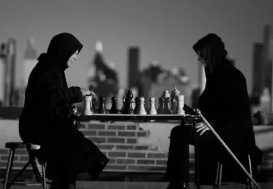 Mitt i en intensiv schackmatch sitter två personer med huvor utomhus på ett tak med stadens silhuett i bakgrunden. Deras koncentration är djup, vilket speglar spelets taktiska natur. Midcent. Underhållning.