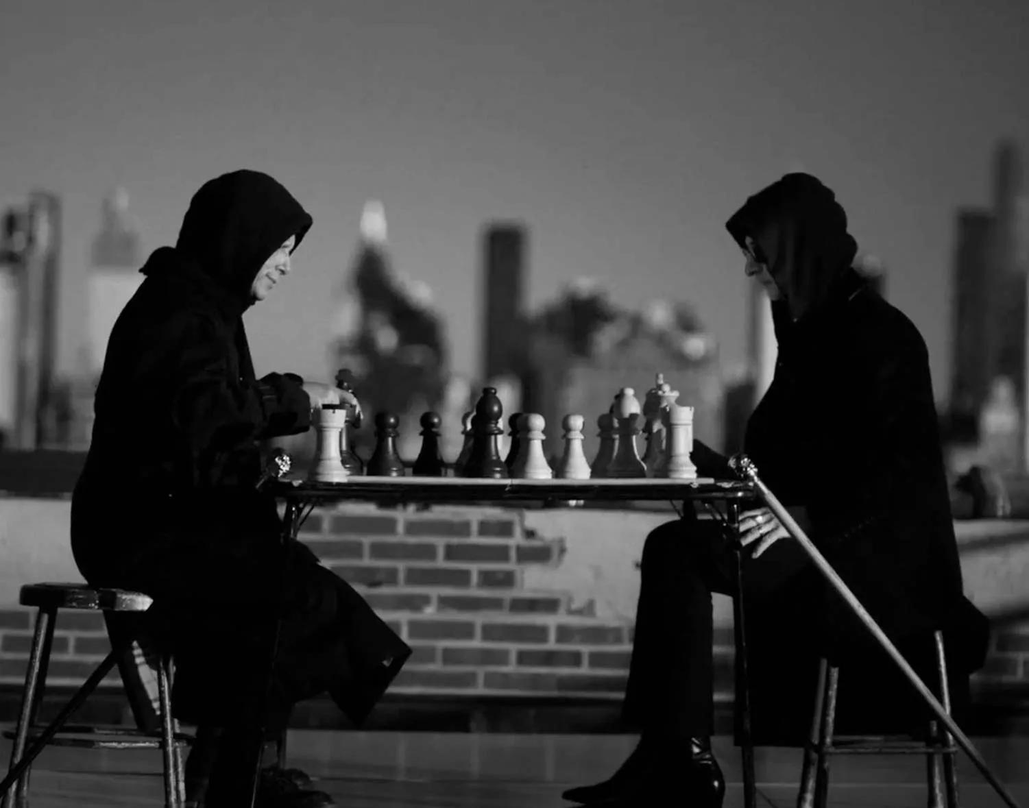 Mitt i en intensiv schackmatch sitter två personer med huvor utomhus på ett tak med stadens silhuett i bakgrunden. Deras koncentration är djup, vilket speglar spelets taktiska natur. Midcent. Underhållning.
