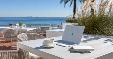 Midcent: En modern bärbar dator, anteckningsbok och kaffekopp på ett soldränkt kafébord vid havet skapar en inspirerande arbetsmiljö. Perfekt för produktivt arbete med drömlik utsikt.Kategori: Resa.