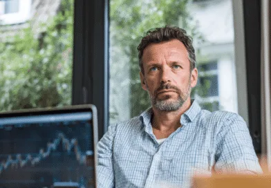 En medelålders man sitter vid ett skrivbord med fokus på en datorskärm som visar aktiemarknadsdata. Bilden illustrerar finansiell analys och investeringsplanering. Midcent. Kategori: Ekonomi.