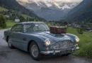 Midcent: En klassisk bil, Aston Martin, står parkerad på en landsväg omgivet av alplandskap. En resväska är placerad på motorhuven. Perfekt för resenärer som söker äventyr med stil. Resa.