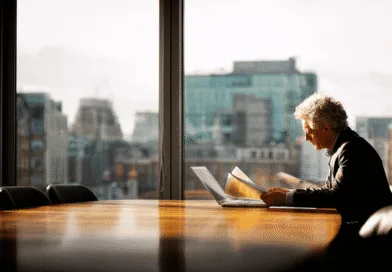Midcent: En medelålders man läser dokument vid ett konferensbord i ett solbelyst kontor med stadsvy. Perfekt för att illustrera affärsstrategi och professionellt beslutsfattande. Ekonomi.