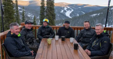 Midcent: En grupp medelålders män sitter på en träveranda i ett snöigt bergslandskap, njutande av varma drycker. Omgivande