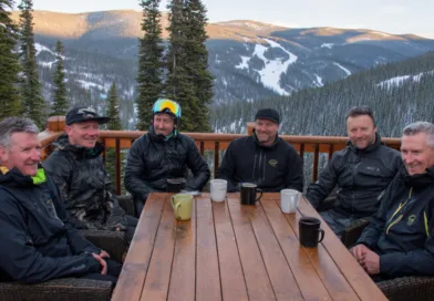 Midcent: En grupp medelålders män sitter på en träveranda i ett snöigt bergslandskap, njutande av varma drycker. Omgivande
