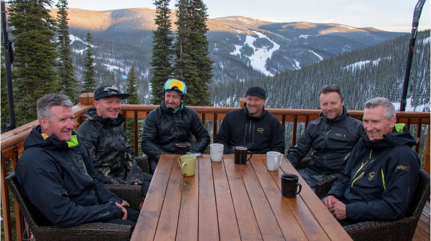 Midcent: En grupp medelålders män sitter på en träveranda i ett snöigt bergslandskap, njutande av varma drycker. Omgivande