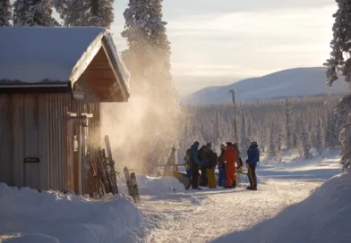 Midcent: En grupp människor njuter av en solig vinterdag vid en fjällstuga. Snötäckta träd och fjälllandskap skapar en
